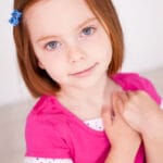 Young girl with straight light brown hair, wearing a pink shirt and blue hair clip, stands indoors with hands over her chest, radiating self-compassion as she looks at the camera.