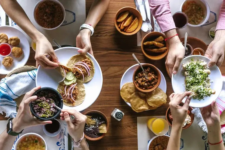 People sharing various Mexican dishes, including tacos, fried plantains, beans, salsas, and guacamole, around a wooden table. Hands are reaching for and serving food.