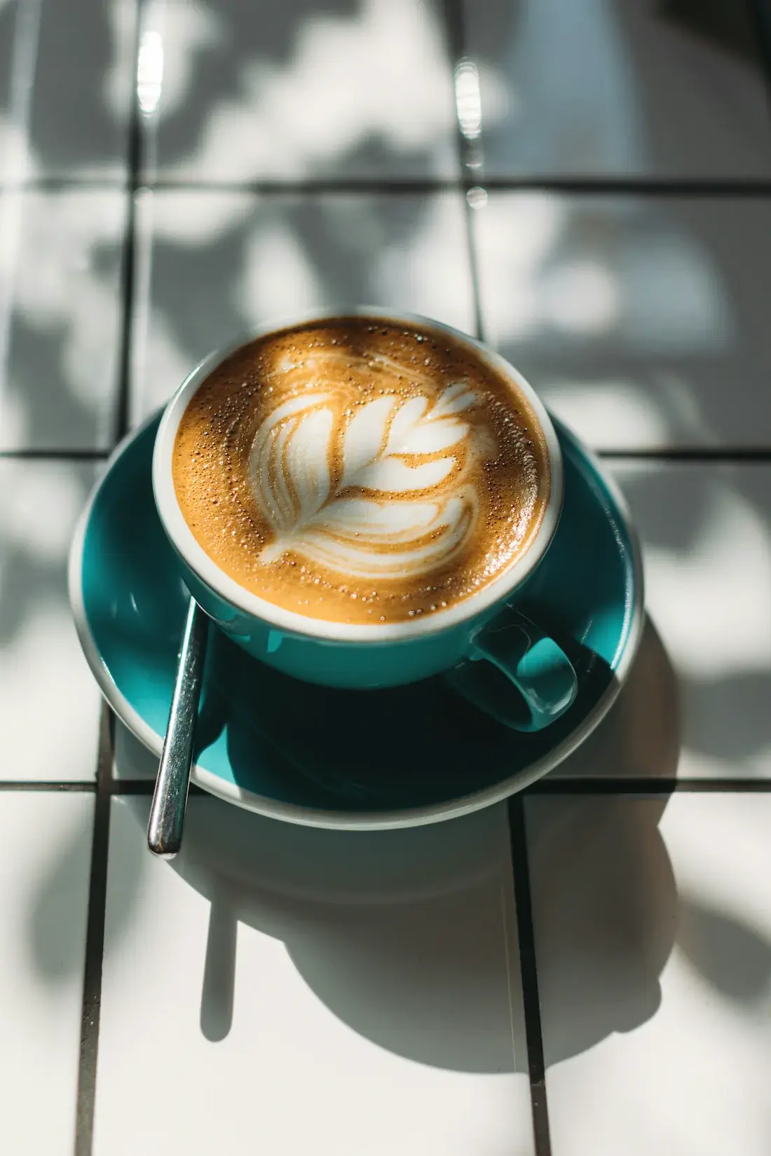 A cup of latte with leaf-shaped latte art sits on a teal saucer with a spoon, placed on a white tiled table with sunlight and shadows.