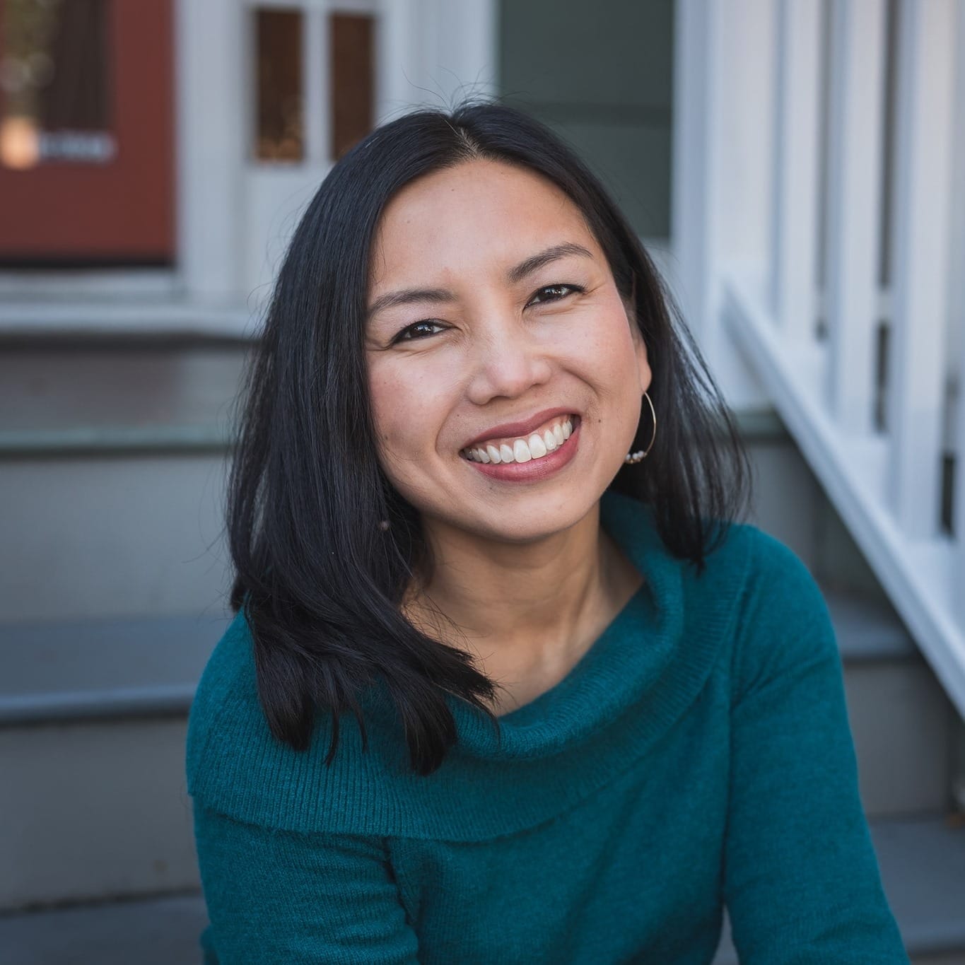 A woman, Minh-Hai Alex, with straight dark hair and hoop earrings, wearing a teal sweater, smiles while sitting on outdoor steps in front of a house.