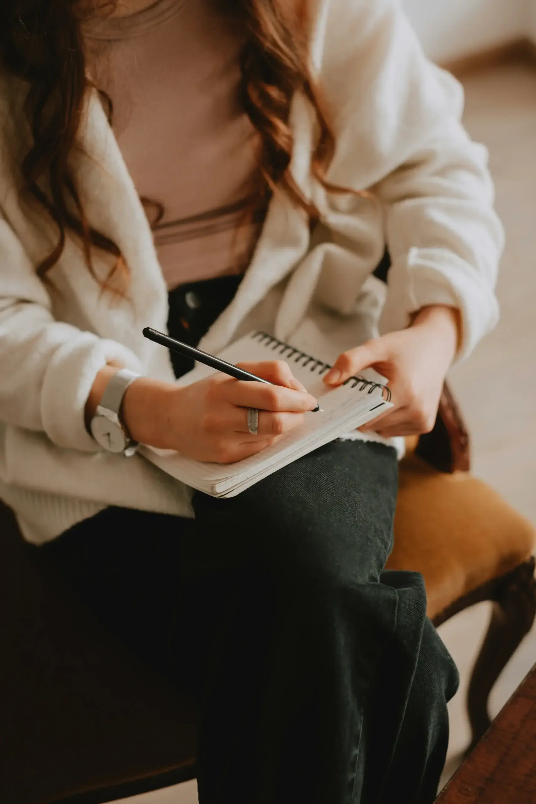 A person wearing a white jacket sits on a chair, writing in a spiral notebook with a pen on their lap.