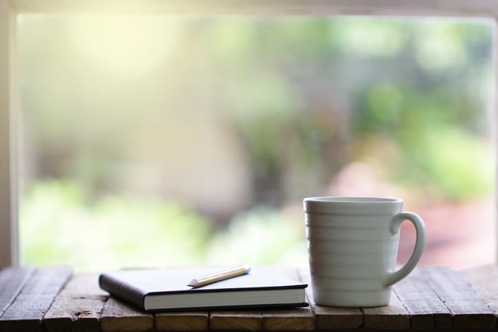 A white coffee mug and a closed notebook with a pen rest on a wooden surface in front of a window with blurred greenery outside.