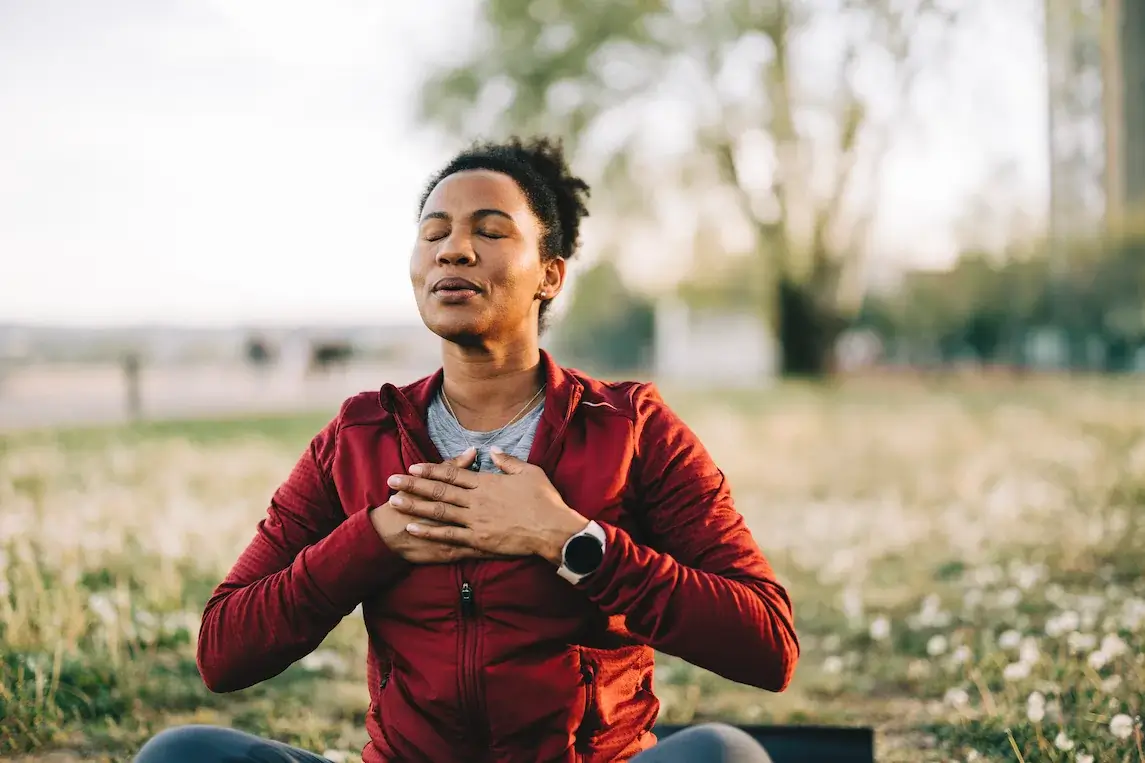 A person in a red jacket sits outdoors in Seattle with eyes closed and hands on chest, appearing to practice deep breathing or iRest Meditation.